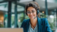 Young Hispanic woman with glasses and headset smiling while working on laptop in modern office space. Customer service representative providing remote support.