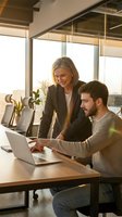 Professional businesswoman mentoring young male colleague at laptop in modern office with warm sunlight streaming through windows during productive meeting.