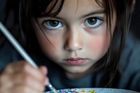 Young girl with intense blue eyes and dark hair focused on drawing or coloring, captured in dramatic lighting that emphasizes facial features and artistic expression.