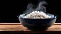 Steaming white rice in dark ceramic bowl on wooden surface against black background, suitable for food and cooking content.