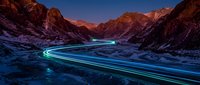Young male driver with light skin tone operating vehicle on winding mountain road at night, long exposure light trails glowing neon blue and green across asphalt, dramatic rocky canyon landscape, dusk sky.
