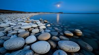 Moonlit coastal landscape with smooth round stones along shoreline reflecting in calm blue water during twilight hour, creating serene natural composition.