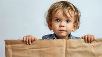 Curious toddler with bright blue eyes peeks over cardboard box edge, wearing denim shirt against white background, expressing wonder and playful innocence.