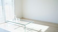 Minimalist glass coffee table with transparent legs in bright, sunlit modern living room with wooden floor and white walls.