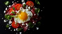 Vibrant breakfast composition with sunny side up egg, fresh cherry tomatoes, prosciutto, feta cheese crumbles and basil leaves on dark background, dramatic lighting.