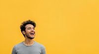 Young man with dark curly hair and beard, appearing to be of Mediterranean or Middle Eastern ethnicity, wearing casual gray t-shirt against bright yellow background. Suitable for lifestyle, portrait, and advertising campaigns.