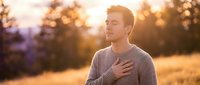 Young Caucasian man in casual gray sweater standing outdoors at golden hour sunset, hand on chest, contemplative expression, peaceful nature background with warm lighting.