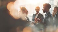 Diverse business professionals engaged in animated conversation at networking event with warm bokeh lighting creating atmospheric corporate ambiance.