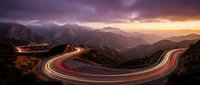 Winding mountain road with light trails at sunset creating dramatic streaks through scenic landscape with purple sky and layered peaks in distance.