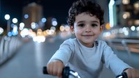 Young boy with curly hair smiling while riding bicycle at night in urban setting with bokeh city lights in background.