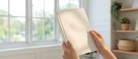 Person holding blank cream colored tablet or clipboard in bright modern living room with large windows, potted plants on shelves, and natural daylight for mockup and branding purposes.