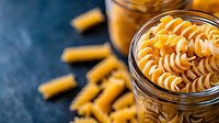 Uncooked golden fusilli pasta in glass jar on dark background with scattered dry macaroni. Fresh ingredients for Italian cuisine and Mediterranean cooking.