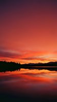 Dramatic sunset over calm lake with silhouetted evergreen forest and mountain range, vibrant orange and red sky reflecting on still water surface for nature and landscape projects.