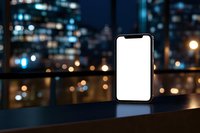 Modern smartphone with blank white screen against blurred urban nightscape with bokeh lights and reflections on dark surface. Mockup ready for design.