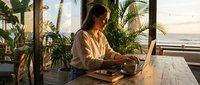 Young woman working on laptop in tropical beachside cafe with palm trees during golden hour sunset creating peaceful remote work atmosphere.