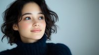 Young Asian woman with natural makeup and short dark wavy hair wearing navy turtleneck sweater looking away with gentle smile against light background.