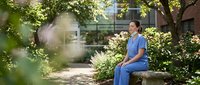 Young Asian female healthcare worker in blue scrubs taking coffee break outdoors in hospital garden courtyard surrounded by blooming flowers and greenery.