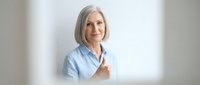 Senior woman with gray hair wearing light blue shirt giving thumbs up gesture against white background, expressing approval and positivity for business and lifestyle content.