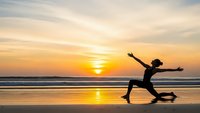 Young woman jumping with joy on beach at golden sunset with arms outstretched against ocean waves and dramatic sky reflecting on wet sand.