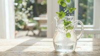 Fresh mint leaves splashing into clear water pitcher on wooden table by sunny window creating refreshing summer drink with natural lighting and bokeh background.
