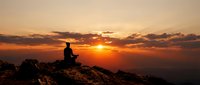 Person silhouette meditating on mountain peak during golden sunset with dramatic clouds and warm orange sky creating peaceful spiritual atmosphere.