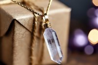 Elegant crystal pendant necklace with gold chain displayed against wrapped gift box with warm bokeh lights creating luxurious jewelry presentation atmosphere.