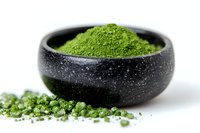Vibrant green matcha powder in black speckled bowl with scattered powder on white surface, ready for beverage preparation and culinary use.