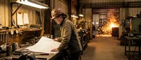 Male architect or engineer in hard hat reviewing blueprints at workbench in industrial workshop with tools, equipment, and warm lighting for construction, design, and professional planning projects.
