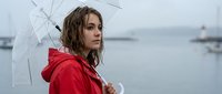Young woman with long brown wavy hair and fair complexion holding transparent umbrella in red jacket by misty harbor with boats, rainy weather mood.