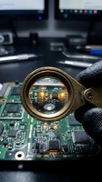 Technician examining computer circuit board with magnifying glass in electronics repair workshop showing detailed microchips and components for diagnostic work.