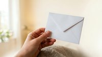 Closeup of hand holding white envelope against blurred interior background with natural light, suitable for mail, communication, and business correspondence concepts.