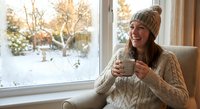 Young woman in cozy knit sweater and beanie enjoying hot drink by window overlooking snowy winter landscape. Warm indoor comfort during cold season.