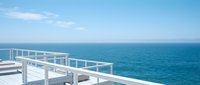 Modern white terrace overlooking turquoise ocean with clear blue sky and fluffy clouds for luxury vacation and travel concepts.