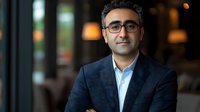 Middle Eastern businessman wearing glasses and navy suit smiles confidently in modern office setting with soft bokeh background lighting, professional corporate portrait.