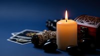 Mystical fortune telling setup with burning candle, tarot cards, sage bundle and wooden chest on dark blue background for spiritual guidance sessions.