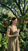 Young woman enjoying refreshing water bottle in lush tropical garden with vibrant orchids and ferns on sunny day for wellness and hydration lifestyle.