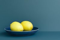 Two fresh yellow lemons resting in a blue ceramic plate against a teal background, minimalist food styling with copy space.