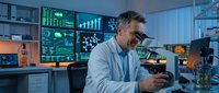 Middle-aged Caucasian male scientist examining sample through microscope in modern laboratory with digital data displays and analytical equipment.