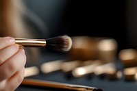 Makeup brush with gold ferrule held in hand against blurred cosmetic products and neutral background, suitable for beauty, cosmetics, and makeup artistry content.