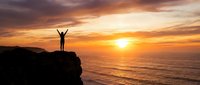 Person with raised arms celebrating success on cliff edge during golden sunset over ocean waves creating inspirational moment of achievement and freedom.