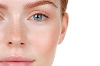 Close-up partial face view of young Caucasian woman with natural makeup, freckles, blue eyes and pink lips showing clean, fresh complexion on white background.