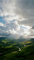 Dramatic sunlight breaks through towering clouds above rolling green hills and winding river valley, creating ethereal rays illuminating the lush landscape below.