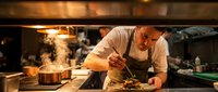 Professional chef plating gourmet dish in commercial kitchen with steam and warm lighting creating culinary atmosphere for restaurant industry content.