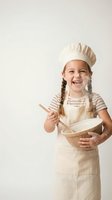 Happy young girl in chef hat and apron mixing ingredients in bowl with wooden spoon on neutral background for cooking education and kitchen activities.