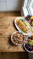 Grilled salmon with herbs and lemon served with quinoa salad and fresh fruit platter on rustic wooden table by window for healthy dining and meal prep.