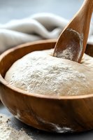 Fresh yeast dough rising in wooden bowl with flour and spoon, macro shot showing soft texture and natural fermentation process for baking bread.