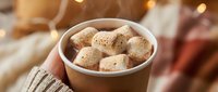 Hot chocolate with marshmallows in hands wearing knitted sweater creating cozy winter atmosphere with warm bokeh lights in background.