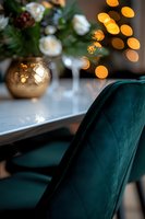 Elegant emerald velvet dining chair with festive golden bokeh lights and white roses in copper vase creating luxurious holiday atmosphere at home.