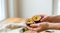 Close-up of hands holding two halves of fresh passion fruit with seeds and pulp visible, isolated on white background for food and culinary design use.
