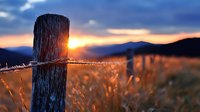Rustic wooden fence post with barbed wire against dramatic sunset sky and golden field, mountains silhouetted in background, warm evening light creating atmospheric rural scene.